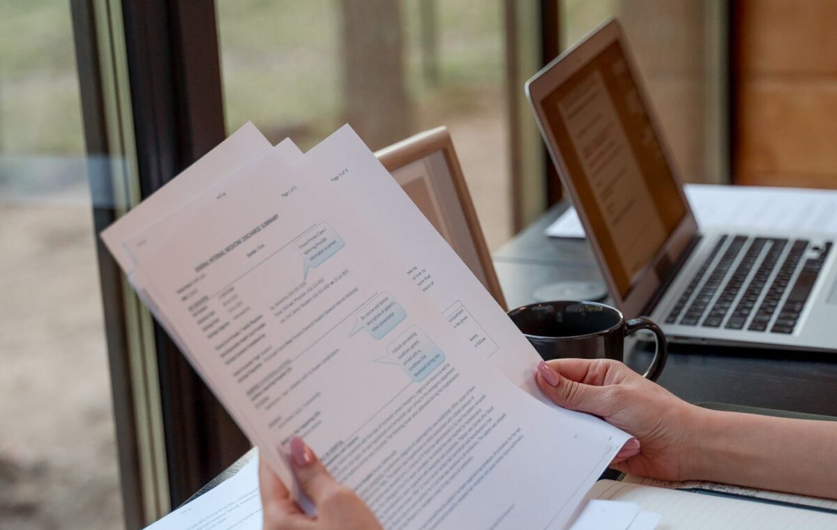Pessoa conferindo documentos diante de um notebook sobre uma mesa