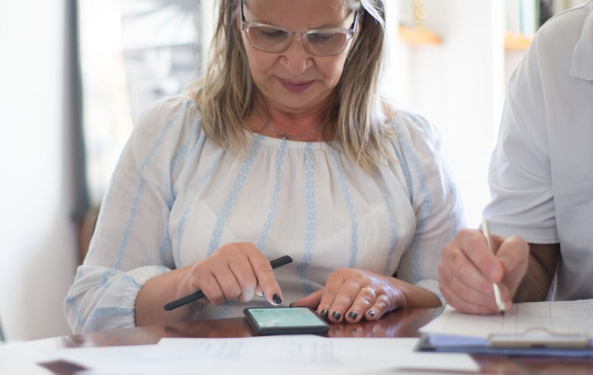 Mulher idosa consulta documentos enquanto usa um smartphone à mesa